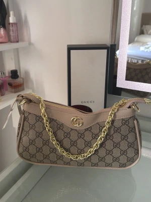  beige väska  - Snygg handväska från  i beige canvas med klassiskt GG-mönster och ljusbruna läderdetaljer. Väskan har en guldfärgad kedja som axelrem och GG-logo framtill. Perfekt accessoar för att lyfta din outfit.