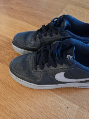 Nike Air Force 1 svarta sneakers - Svarta Nike Air Force 1 sneakers med vit swoosh och vit sula. Klassisk låg modell med perforerad tå och snörning. Skorna har en robust design och är tillverkade i läder och textil. Perfekt för dig som gillar streetstyle och ikoniska sneakers. 
