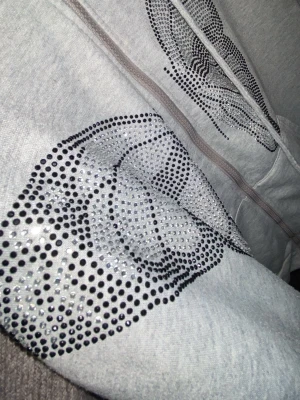 Grå hoodie med strassmönster - Snygg grå hoodie med dragkedja och huva, dekorerad med ett coolt mönster av svarta och silvriga strasstenar på framsidan. Perfekt för dig som gillar lite extra bling i vardagen. Mjuk bomullsblandning och dragsko i huvan.