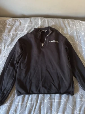 Svart Calvin Klein halvzip tröja - Svart långärmad tröja från Calvin Klein med halvzip och vit logga på bröstet. Tröjan har en hög krage och är tillverkad i mjukt material. På baksidan finns en vit patch med texten 'JEANS' nere vid fållen.