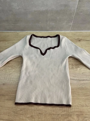 Beige ribbad topp med brun kant - Trendig ribbad topp i beige med mörkbruna kanter och vågig v-ringning. Långärmad och figurnära passform, perfekt att styla med jeans eller kjol. Materialet känns mjukt och stretchigt.