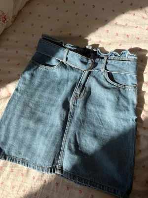 Blå jeanskjol med bälte - Snygg blå jeanskjol i kort modell med hög midja och tillhörande bälte. Kjolen har klassiska fickor fram och bak samt en vågig kant upptill för en trendig look.