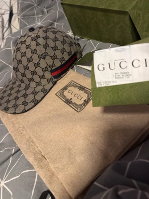 Gucci keps med GG-mönster - Säljer en snygg Gucci keps i beige med klassiskt GG-monogram över hela kepsen. Kepsen har en röd och marinblå rand på sidan och är tillverkad i canvasmaterial. Perfekt accessoar för dig som gillar exklusiv och trendig streetwear. Den är helt ny köptes 2023 men aldrig använd kvitto finns o kartong me påse osv den sälj int längre 