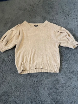 Beige stickad tröja från Lindex - Snygg beige stickad tröja från Lindex med puffärmar och ribbade muddar. Tröjan har rund halsringning och kortare ärmar med volym, vilket ger en trendig och avslappnad look. Perfekt att matcha med jeans eller kjol för en cool vardagsstil.