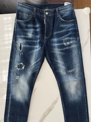 Dsquared2 Jeans - Säljer ett par blå Dsquared2 jeans med distressed detaljer och slitningar på både fram och baksida. Jeansen är slimfit och i ny skick. Jag använde de en gång men de var inte min typ av jeans. Priset kan diskuteras.