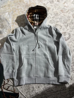 Grå zip-hoodie från Burberry - Snygg grå hoodie med dragkedja från Burberry. Klassisk huva med det ikoniska rutmönstret på insidan och broderad logga på bröstet. Mjuk bomullsblandning, ribbade muddar och fickor framtill. Perfekt för en chill och stilren look.