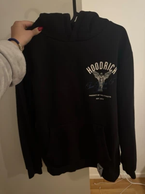 Svart hoodie från Hoodrich - Svart hoodie från Hoodrich med tryck av en ängel och texten 'From Nothing To Something' på ryggen. Framsidan har Hoodrich-logga och mindre tryck. Klassisk känguruficka och huva. Perfekt för dig som gillar streetwear och snygga detaljer.