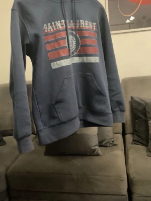 Blå Saint Laurent hoodie med tryck - Snygg blå hoodie från Saint Laurent med vit och röd logga och tryck framtill. Klassisk känguruficka och justerbar huva med dragsko. Perfekt för en avslappnad och trendig stil. Passar med med allt och är perfekt nu till våren och sommarkvällar. Den är knappt använd. Hör av dig för fler bilder och diskussion av pris