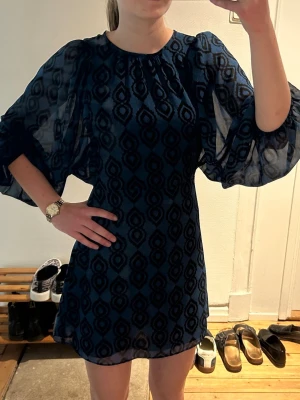 Blå mönstrad kortklänning med puffärm - Kort klänning i blått med svart geometriskt mönster och transparenta puffärmar. Klänningen har rund halsringning och är tillverkad i ett lätt, luftig material som ger en cool vibe. Perfekt för dig som gillar statement-ärmar och unik stil.