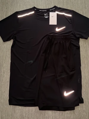 Svart träningsset från Nike - Snyggt svart träningsset från Nike med t-shirt och shorts. Båda plaggen har reflekterande vita Nike-loggor och är gjorda i ett lätt, snabbtorkande funktionsmaterial. Perfekt för löpning eller gym. Tidlös design med rund hals och elastisk midja på shortsen.                                       ⚠️ STORLEKSINFO: Lappen säger L (Kina), men motsvarar en EU Small (S). • Bröst: 95–101 cm. • Midja: 88–94 cm. Först till kvarn! 🚀 