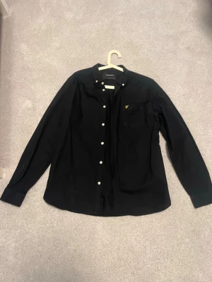 Svart skjorta från Lyle & Scott - Stilren svart skjorta från Lyle & Scott med klassisk krage, vita knappar och gul logga på bröstfickan. Skjortan har lång ärm och passar perfekt till en trendig look. Tillverkad i mjukt bomullstyg för skön känsla.