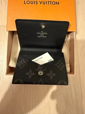 Louis Vuitton svart monogram plånbok - Snygg svart plånbok från Louis Vuitton i klassiskt monogrammönster. Plånboken är tillverkad i tåligt skinn och har tryckknappsstängning. Perfekt storlek för kort och sedlar, med stilren design och exklusiv känsla.