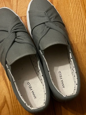 Grå slip-on sneakers Anna Field - Snygga grå slip-on sneakers från Anna Field med twistad detalj på ovandelen. Skorna har vit sula och innersula med prickigt mönster. Tillverkade i textilmaterial och har en låg profil som passar till många olika stilar.