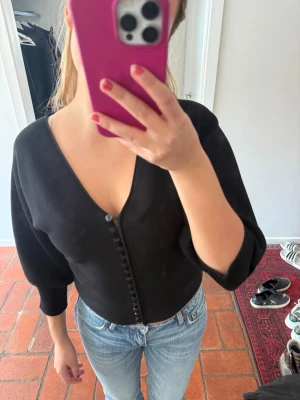 Svart croppad blus med knappar - Svart croppad blus med v-ringning och dekorativa knappar framtill. Modellen har trekvartsärmar och en figurnära passform som ger en snygg siluett. Perfekt att styla med jeans eller kjol för en trendig look.