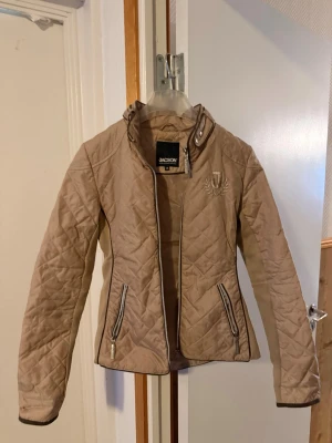 Beige quiltad vindjacka från Jacson - Snygg beige vindjacka från Jacson med quiltat mönster och smal passform. Jackan har hög krage med spänne, två dragkedjefickor och ett broderat emblem på bröstet. Materialet är lätt och passar perfekt för blåsiga dagar. Nypris är cirka 800 kr säljer min för 200