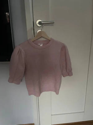 Ljusrosa stickad topp med puffärm - Supersöt ljusrosa stickad topp med puffärmar och rund hals. Tröjan har kortare ärmar med ribbade muddar och en mjuk, skön känsla. Perfekt för dig som gillar en feminin och trendig stil. Materialet känns mjukt och stretchigt. Egentligen barn storlek 158/164 men passar en xs
