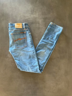 Blå jeans från Nudie Jeans - Snygga blå jeans från Nudie Jeans med klassisk femficksdesign och orangea detaljer på bakfickan. Jeansen har en rak passform och är tillverkade i mjukt denimtyg. Perfekta för en avslappnad och trendig stil.     Finns en liten lagring på dem, ser inget av lagringen 