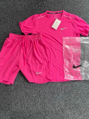 Nike Mliler Rosa Set - Nike träningsset med t-shirt och shorts i neonrosa. Setet har en sportig design med silverfärgade Nike-loggor på både tröjan och shortsen. Tillverkat i lätt och snabbtorkande material som passar perfekt för träning och aktivitet. M/ S storlek