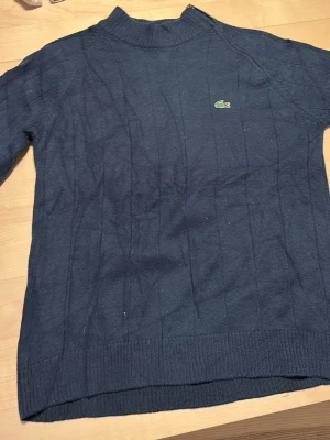 Mörkblå stickad tröja från Lacoste - Mörkblå stickad tröja från Lacoste med diskret strukturmönster och klassisk krokodillogga på bröstet. Tröjan har hög krage och ribbade muddar vid ärmslut och nederkant. Pris kan diskuteras