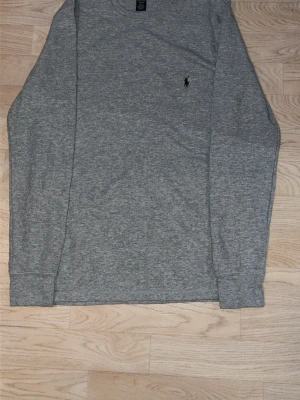 Grå långärmad loungewear från Polo Ralph Lauren - Mysig grå långärmad loungewear-tröja från Polo Ralph Lauren. Tröjan har rund halsringning, ribbade muddar och diskret broderad logga på bröstet. Perfekt för chill dagar hemma. Tillverkad i mjukt, ribbat material för extra komfort.
