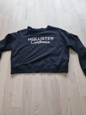 Svart Hollister California hoodie - Svart hoodie från Hollister med vit och rosa text 'Hollister California' på bröstet. Klassisk rak modell med ribbade muddar och rund halsringning. Perfekt för en chill och avslappnad stil. Materialet känns mjukt och skönt mot huden.