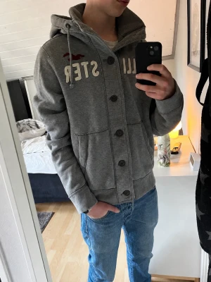  Vintage Hollister Cardigan hoodie med knappar - Säljer nu denna snygga grå vintage  Hollister cardigan. Tröjan har vit Hollister text broderat på framsidan, samt en vinröd Hollister logga. Står att tröjan är i storlek XL, men passar som L/M.  Skick 9/10.  Modellen på bilden är 183 och väger ca 68 kilo.  Längd: 68 cm Bredd (armhåla till armhåla): 55 cm Skriv gärna om ni har några mer funderingar🙂