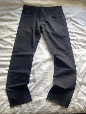 Svarta chinos från Hugo Boss - Snygga svarta chinos från Hugo Boss med stretch passform och fem fickor. Byxorna har diskreta detaljer och är tillverkade i ett mjukt bomullsmaterial med lite stretch för extra komfort. Perfekta för en stilren look.