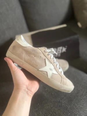 Beige Golden Goose Superstar sneakers - Snygga beige Golden Goose Superstar sneakers i mocka med vit stjärndetalj på sidan och vita skosnören. Skorna har en klassisk låg siluett och vit sula, samt diskreta guldfärgade detaljer. Perfekta för en trendig och avslappnad look.