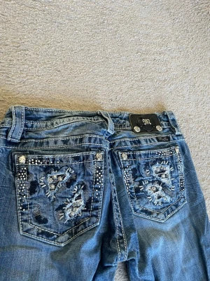 Miss Me blå bootcut jeans med nitar - Snygga blå bootcut jeans från Miss Me med slitningar och dekorerade bakfickor. Fickorna har broderier, nitar och strassdetaljer som ger ett unikt och trendigt intryck. Klassisk femficksmodell i denim med låg midja och kontrastsömmar.