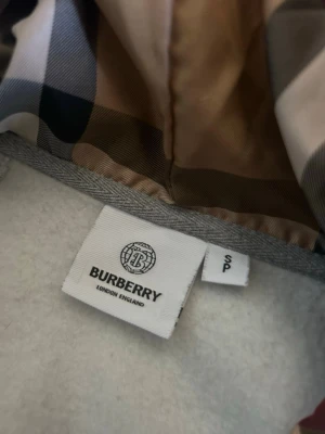 Grå hoodie från Burberry med dragkedja - Snygg grå hoodie från Burberry med klassiskt rutigt foder i huvan. Tröjan har dragkedja framtill, två fickor och ribbade muddar. Perfekt för dig som vill ha en stilren och exklusiv look med en touch av ikoniskt mönster. Pris går att diskutera 