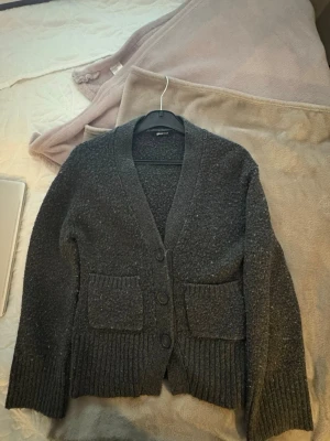 Mörkgrå kofta med fickor - Mysig mörkgrå kofta från Gina Tricot med grovstickad känsla. Koftan har djup V-ringning, tre knappar framtill och två stora fickor. Ribbstickade muddar vid ärmslut och nederkant ger en avslappnad look