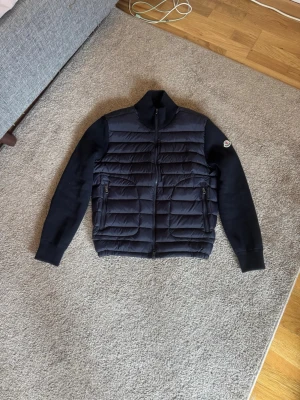 Mörkblå pufferjacka från Moncler - Snygg mörkblå pufferjacka från Moncler med quiltad front och stickade ärmar. Jackan har dragkedja framtill, två fickor med dragkedja och Moncler-logga på vänster ärm. Perfekt för kalla dagar och riktigt trendig stil. Vid snabb affär kan priset diskuteras.