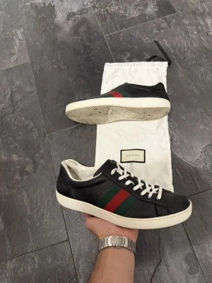 Gucci svarta sneakers med ränder - Snygga svarta sneakers från Gucci med klassiska gröna och röda ränder på sidan. Skorna har vita snören och vit sula, samt en låg profil. Tillverkade i läder och levereras med original dustbag.
