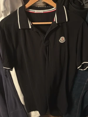 Svart Moncler pikétröja med vit kant - Svart pikétröja från Moncler med klassisk krage och vita ränder längs krage och ärmslut. Tröjan har Moncler-logga på bröstet och korta ärmar. Perfekt för en stilren och sportig look.
