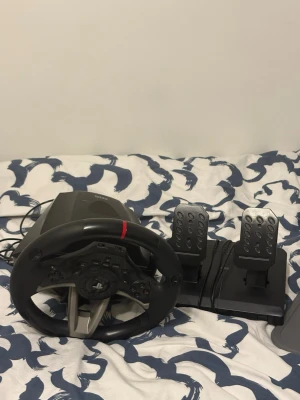  En PS ratt  - Säljer en Ps ratt  racingratt med tillhörande pedaler. Ratten är svart med detaljer i silver och har en röd markering upptill.       Den har används i 3 till 4 månader funkar till Ps och Pc