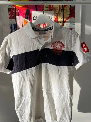 Vit och marinblå piké från Peak Performance - Klassisk pikétröja från Peak Performance i vitt med bred marinblå rand över bröstet. Röd brodyr på bröstet och siffran 86 på ärmen. Krage med randig detalj på insidan och knäppning framtill. Perfekt för en sportig och stilren look.