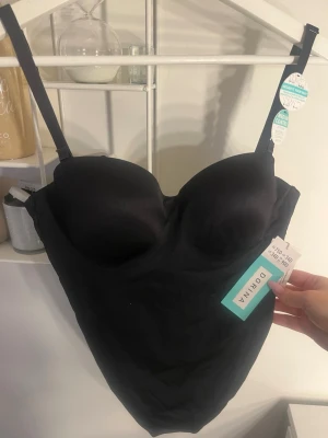 Svart shapingbody från Dorina - Snygg svart shapingbody från Dorina med vadderade kupor och justerbara axelband. Tillverkad i mjukt och stretchigt material för en bekväm passform. Perfekt under tighta plagg för en slät siluett. Klassisk design med diskret look.