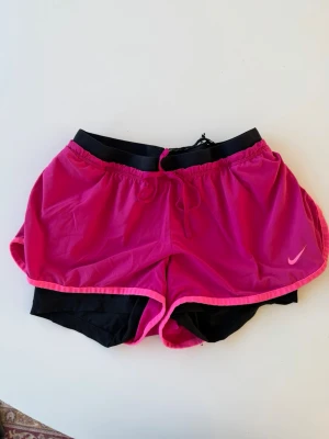 Rosa löparshorts från Nike - Snygga rosa löparshorts från Nike med svarta innershorts och elastisk midja. Shortsen har en liten skada (se bilder) därav billigt pris 