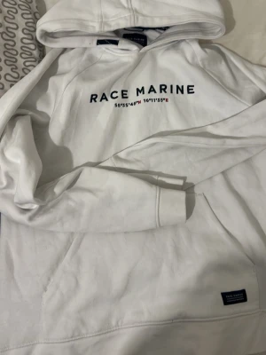 Vit hoodie från Race Marine - Snygg vit hoodie från Race Marine med tryck på bröstet och en liten patch vid fickan. Klassisk känguruficka och huva. Perfekt för en chill och stilren look. Mjuk bomullskänsla och relaxed fit. Köpte den dyr men vill sälja den för den var liten på mig 