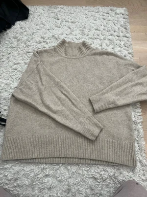Beige stickad tröja med hög krage - Beige stickad tröja med hög ribbad krage och breda ribbade muddar vid ärmslut och nederkant. Tröjan har en avslappnad passform och är tillverkad i ett mjukt material som känns skönt mot huden. Perfekt för lager-på-lager och enkel att matcha.
