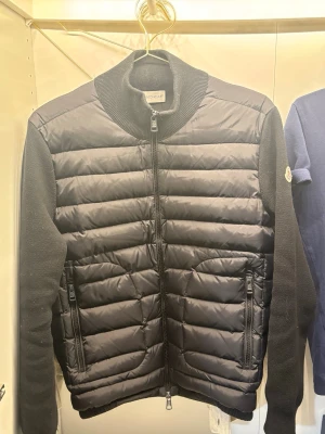 Moncler cardigan - Snygg svart pufferjacka från Moncler med quiltad front och rygg i glansigt material samt stickade ärmar och krage. Jackan har två dragkedjefickor framtill och Moncler-logga på ärmen. Perfekt för kalla vinterdagar och riktigt trendig stil.