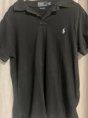 Svart pikétröja från Ralph Lauren - Klassisk svart pikétröja från Polo Ralph Lauren med vit broderad logga på bröstet. Tröjan har korta ärmar, krage och knappar vid halsen. Tillverkad i ett mjukt och slitstarkt bomullsmaterial med custom fit-passform.