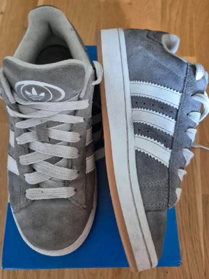 Grå Adidas campus - Snygga grå Adidas campus med klassiska vita ränder på sidorna och vit sula. Använda men fortfarande väldigt fina. 
