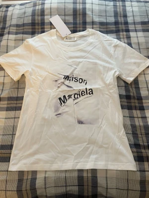 Vit Maison Margiela t-shirt - Vit t-shirt från Maison Margiela med tryckt grafiskt motiv och text på framsidan. Klassisk passform med rund hals och korta ärmar. Tillverkad i mjuk bomull, perfekt för en clean och stilren look.