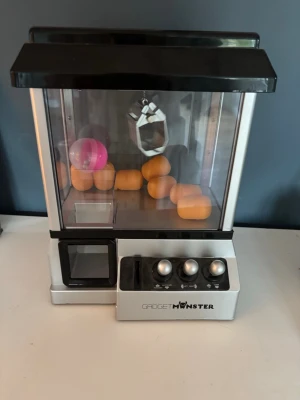 Claw machine från Gadget Monster - Rolig claw machine i silver och svart från Gadget Monster. Maskinen har tre spakar för att styra klon och en transparent plastkupa där man ser leksakerna inuti. Perfekt för att utmana vänner på att plocka upp småprylar eller godis.