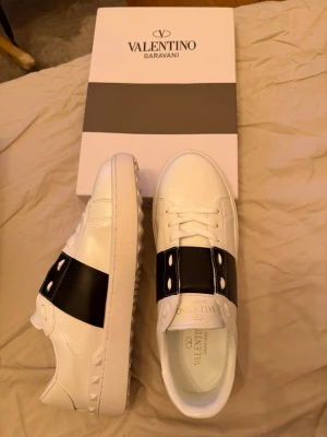 Valentino Garavani vita sneakers - Stilrena vita sneakers från Valentino Garavani med bred svart läderdetalj över vristen och ikoniska nitar längs sulkanten. Skorna har klassisk låg profil, vita snören och en robust gummisula. Perfekta för dig som gillar lyxig och modern streetstyle. Helt nya oanvända, säljer pga att dem är för små.