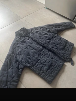 Svart quiltad pufferjacka - Säljer en svart quiltad pufferjacka med hög krage och två stora framfickor. Jackan har dragkedja framtill och ett mönstrat yttertyg med vågformade sömmar. Perfekt för kalla dagar och riktigt skön att bära.