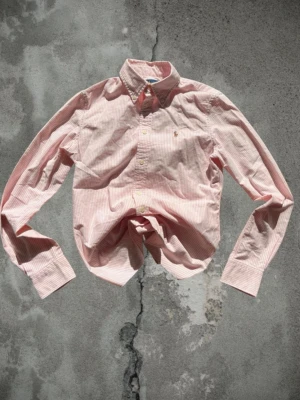 Rosa och vit randig skjorta från Polo Ralph Lauren - Klassisk rosa och vit randig skjorta från Polo Ralph Lauren med broderad logga på bröstet. Skjortan har lång ärm, knappar framtill och är tillverkad i mjuk bomull. Perfekt för en stilren look.