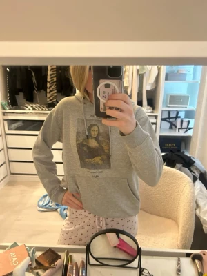 Grå hoodie med tryck framtill - Snygg grå hoodie från A.P.O.H med ett konstmotiv tryckt på bröstet. Klassisk modell med huva och dragsko, samt ribbade muddar vid ärmslut och nederkant. Perfekt för en chill och avslappnad stil. Mona Liza ifram . Nypris är ish 600kr