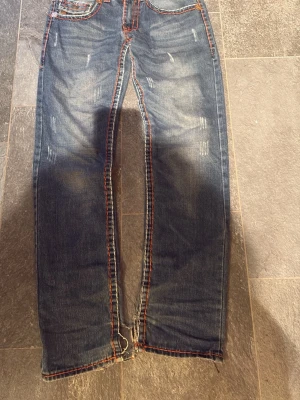 True religion  - Säljer ett par blå jeans med slitningar och tydliga orange kontrastsömmar. Jeansen har raka ben, fem fickor och bakfickor med dekorativa sömmar och knappar. Materialet är klassisk denim och passformen är relaxed.